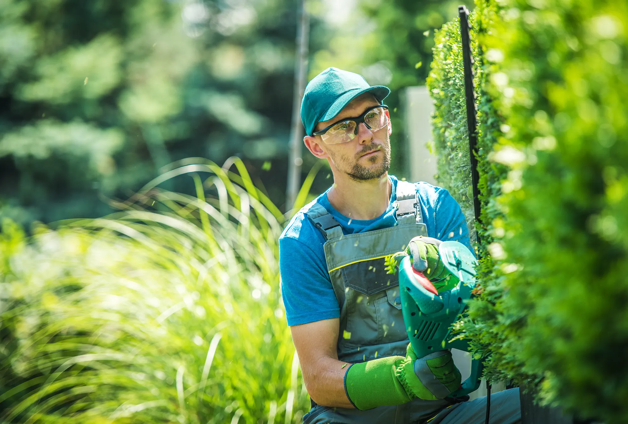 Travailleur en uniforme effectuant la taille de haie avec un taille-haie électrique, dans un jardin résidentiel verdoyant.